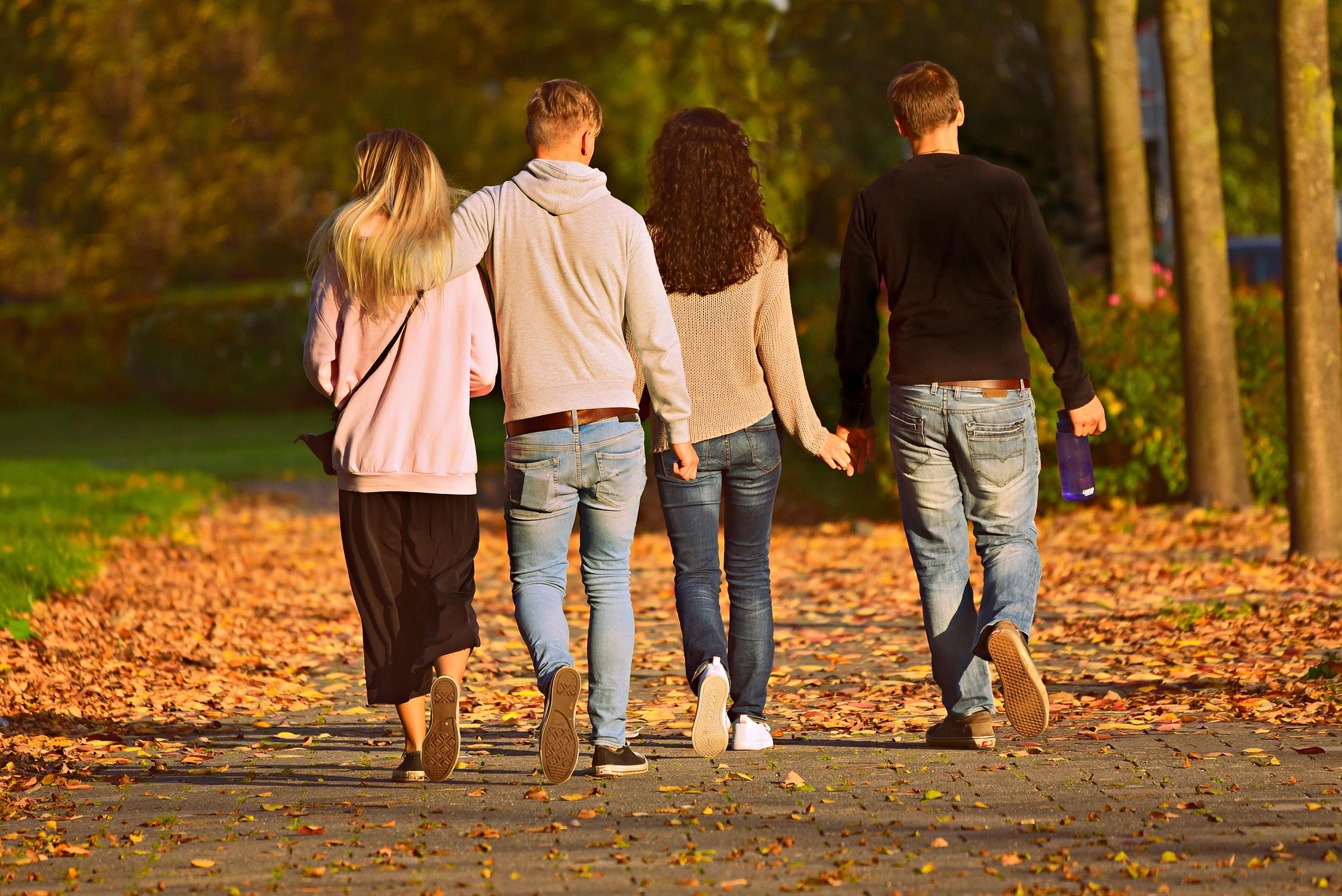 Two couples walking hand in hand along a path in a park with autumn leaves.